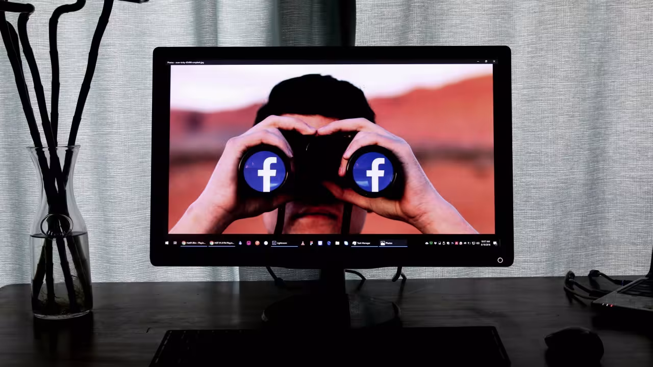 Na ekranie monitora postać z twarzą zasłoniętą lornetką z białymi literami f na niebieskim tle (logo Facebooka) na obiektywach