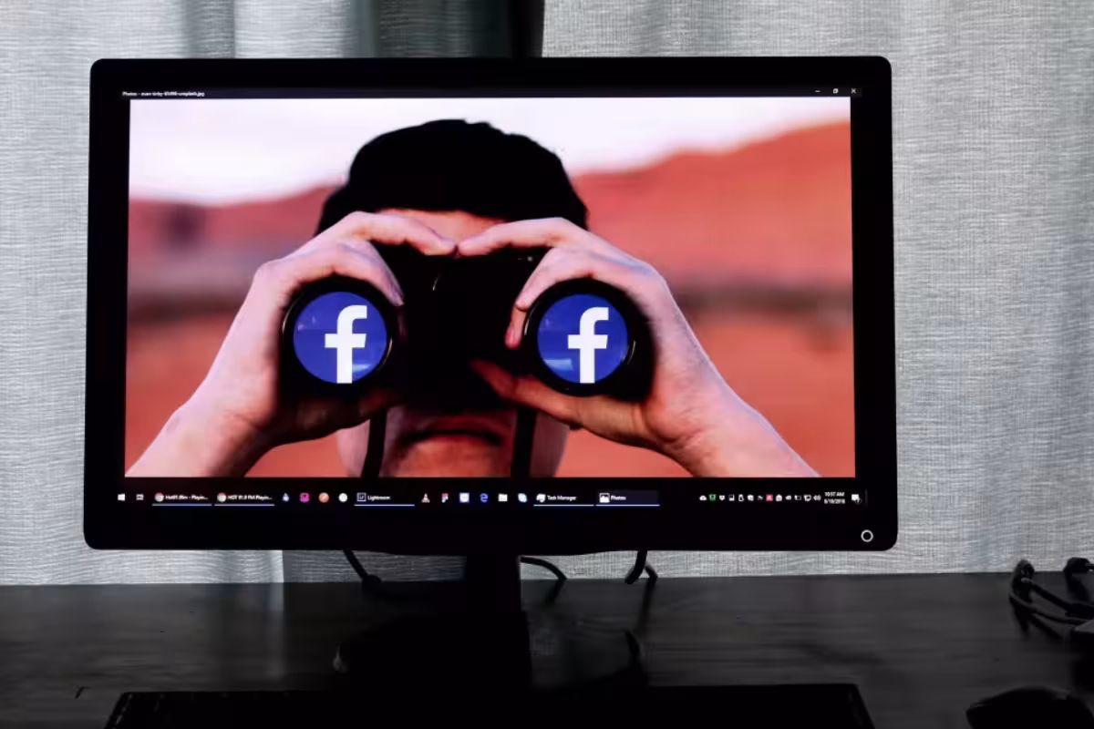 Na ekranie monitora postać z twarzą zasłoniętą lornetką z białymi literami f na niebieskim tle (logo Facebooka) na obiektywach