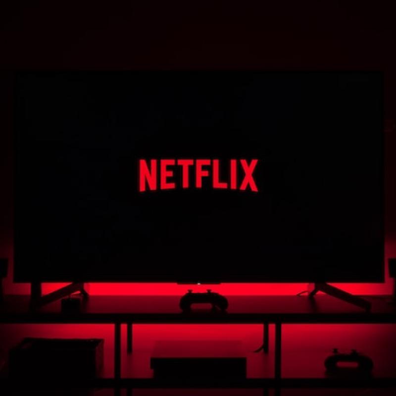 Telewizor z wyświetlonym logiem "Netflix" na czarnym tle. Przed monitorem czerwone krzesła