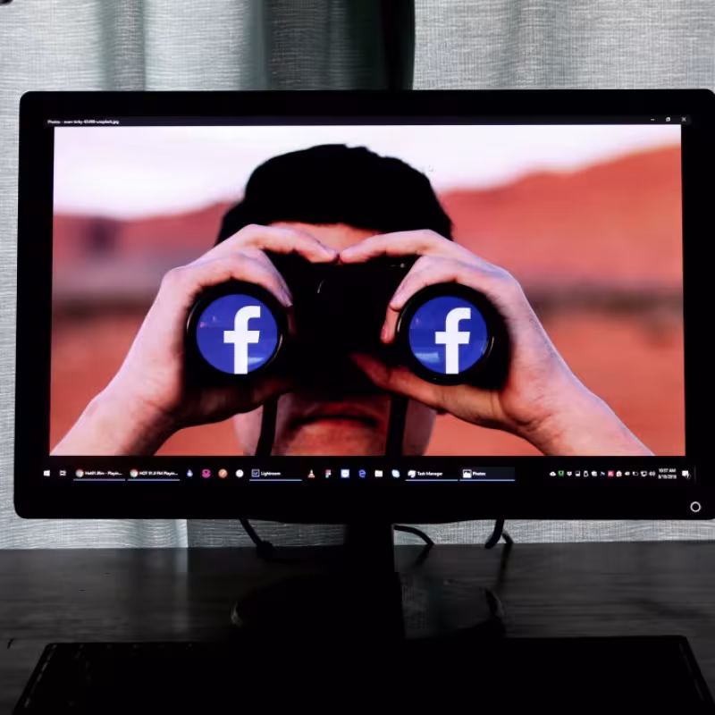 Na ekranie monitora postać z twarzą zasłoniętą lornetką z białymi literami f na niebieskim tle (logo Facebooka) na obiektywach