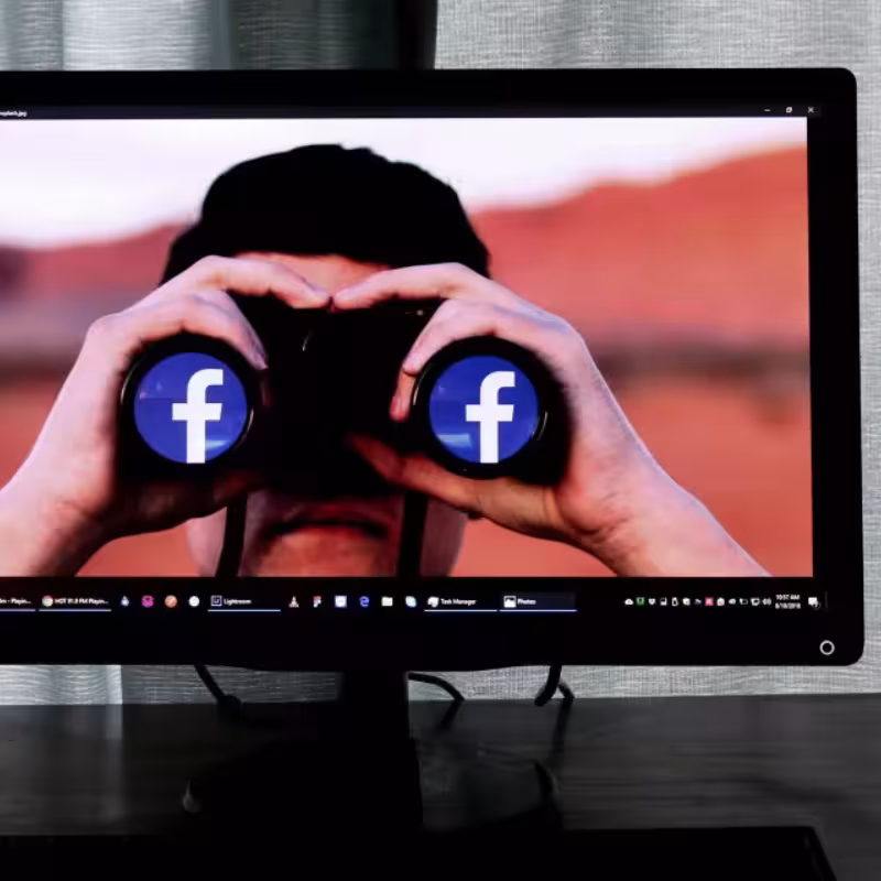 Na ekranie monitora postać z twarzą zasłoniętą lornetką z białymi literami f na niebieskim tle (logo Facebooka) na obiektywach